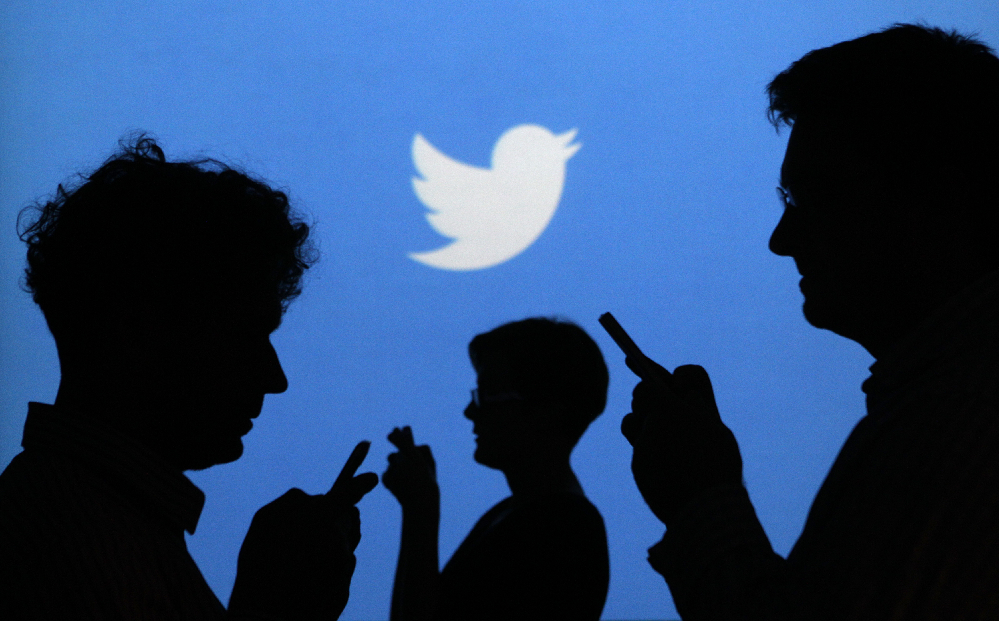 People holding mobile phones are silhouetted against a backdrop projected with the Twitter logo