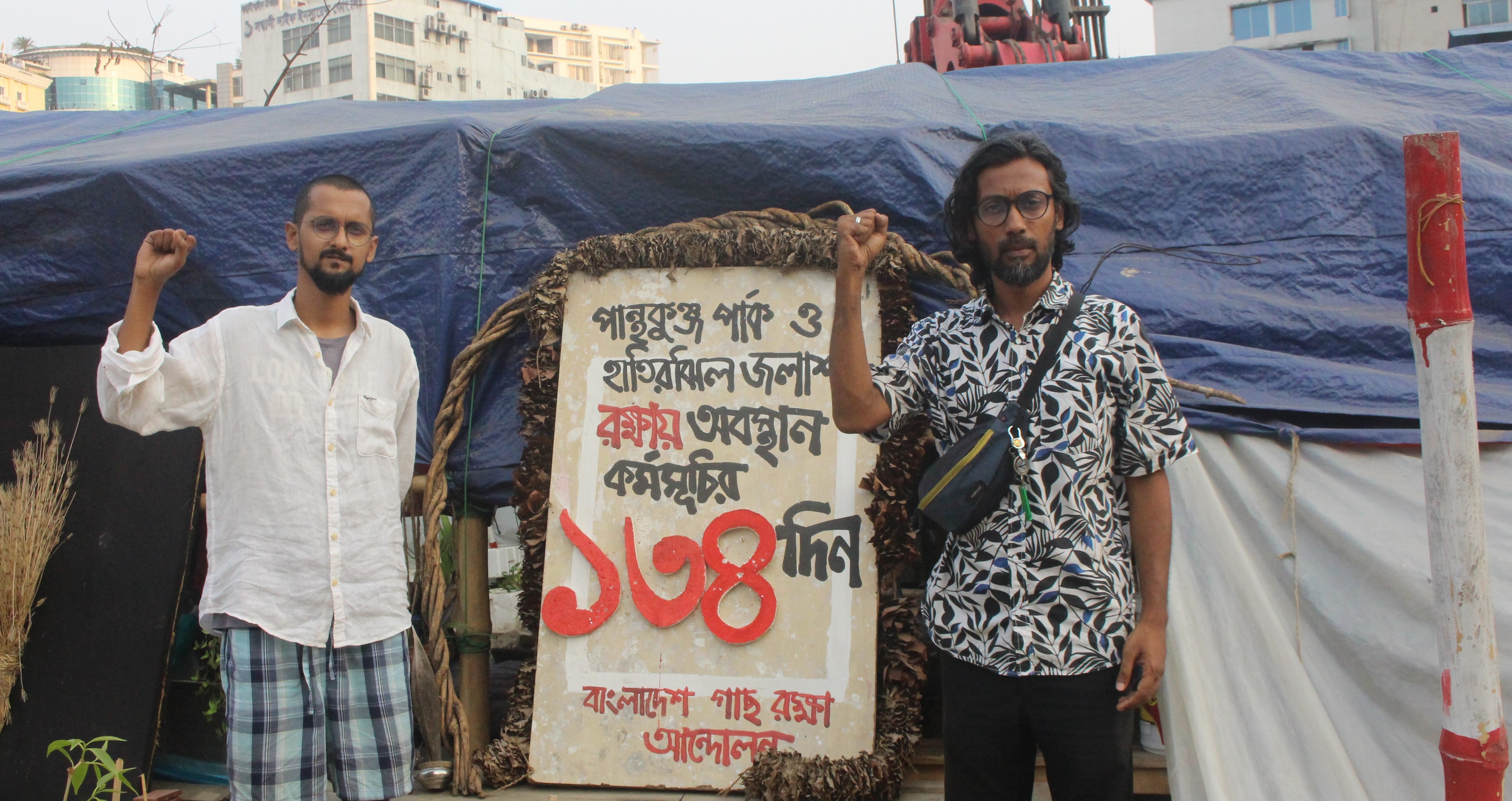 Amirul Rajiv and Naim Ul Hasan, two coordinators of Bangladesh Tree Protection Movement, pose before the makeshift tent where they have been staging a sit-in for more than four months, Dhaka, Bangladesh, April 26, 2025. Md. Tahmid Zami/ Thomson Reuters Foundation